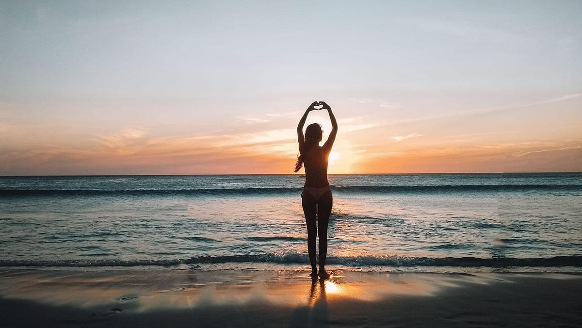 Silueta de una mujer formando un corazón con sus manos sobre su cabeza en la Playa do Grande en Arraial do Cabo, Rio de Janeiro, durante un hermoso atardecer con cielo de tonos cálidos y reflejos dorados sobre las olas suaves.