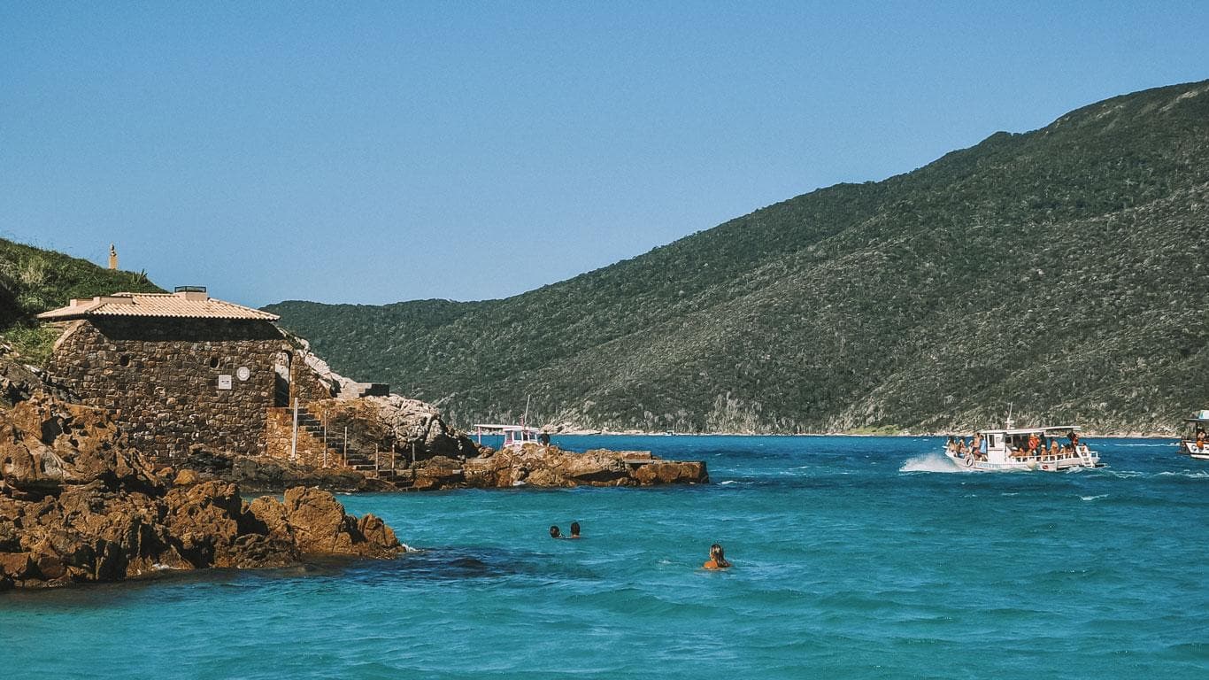Fotografía de una playa en Arraial do Cabo con aguas cristalinas azul turquesa y bañistas disfrutando del día. A la izquierda, una estructura de piedra con techo de paja frente a colinas cubiertas de vegetación, mientras un barco lleno de turistas navega a la derecha.