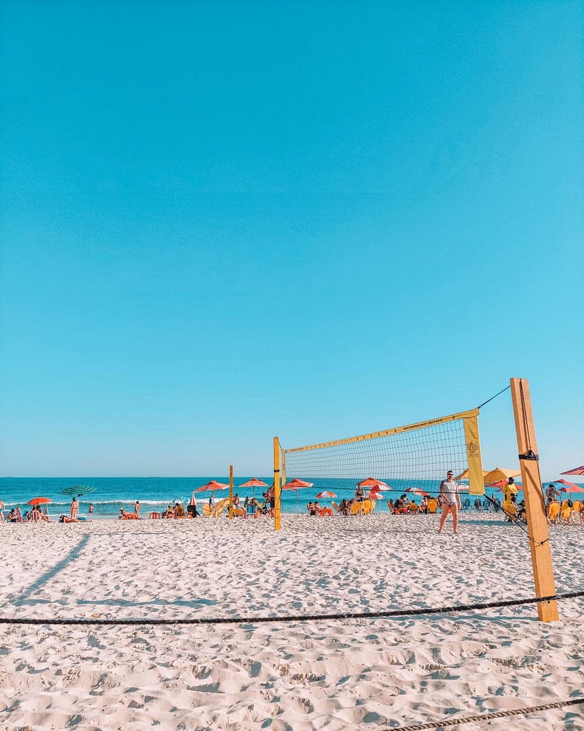Una escena vibrante en la playa de Barra da Tijuca en Río de Janeiro, con una red de voleibol playero en primer plano, el cielo azul claro arriba y personas relajándose bajo sombrillas naranjas en la arena blanca cerca del océano en enero, el mejor mes para visitar Río de Janeiro.